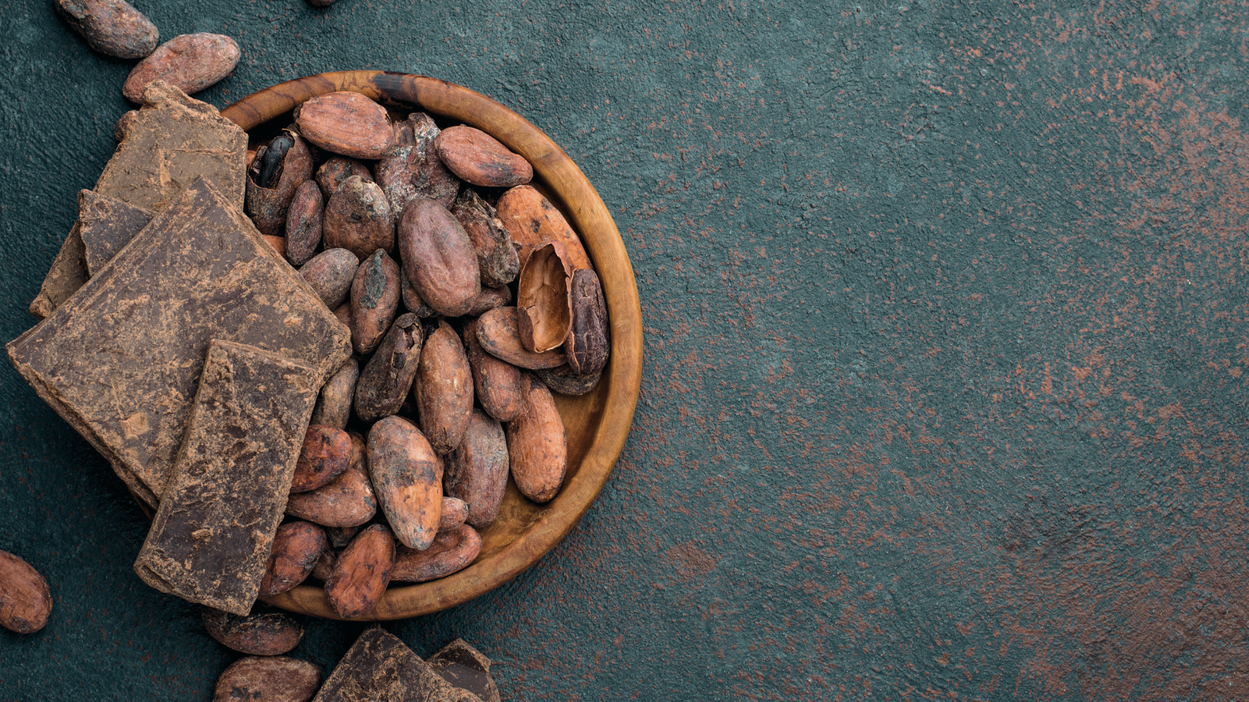 Top-down view of a wooden bowl filled with cacao beans and cacao paste chunks on a textured dark countertop, illustrating raw cacao ingredients.