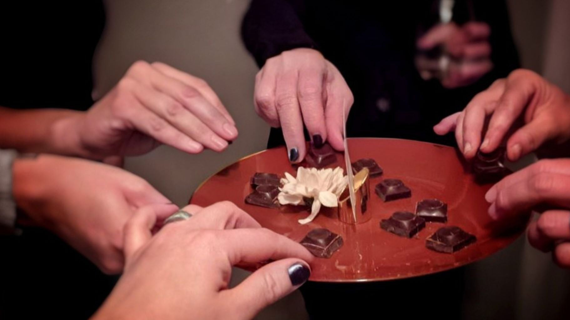 Craft chocolate tasting with hands reaching for dark chocolate squares on a copper platter with a white flower centerpiece.