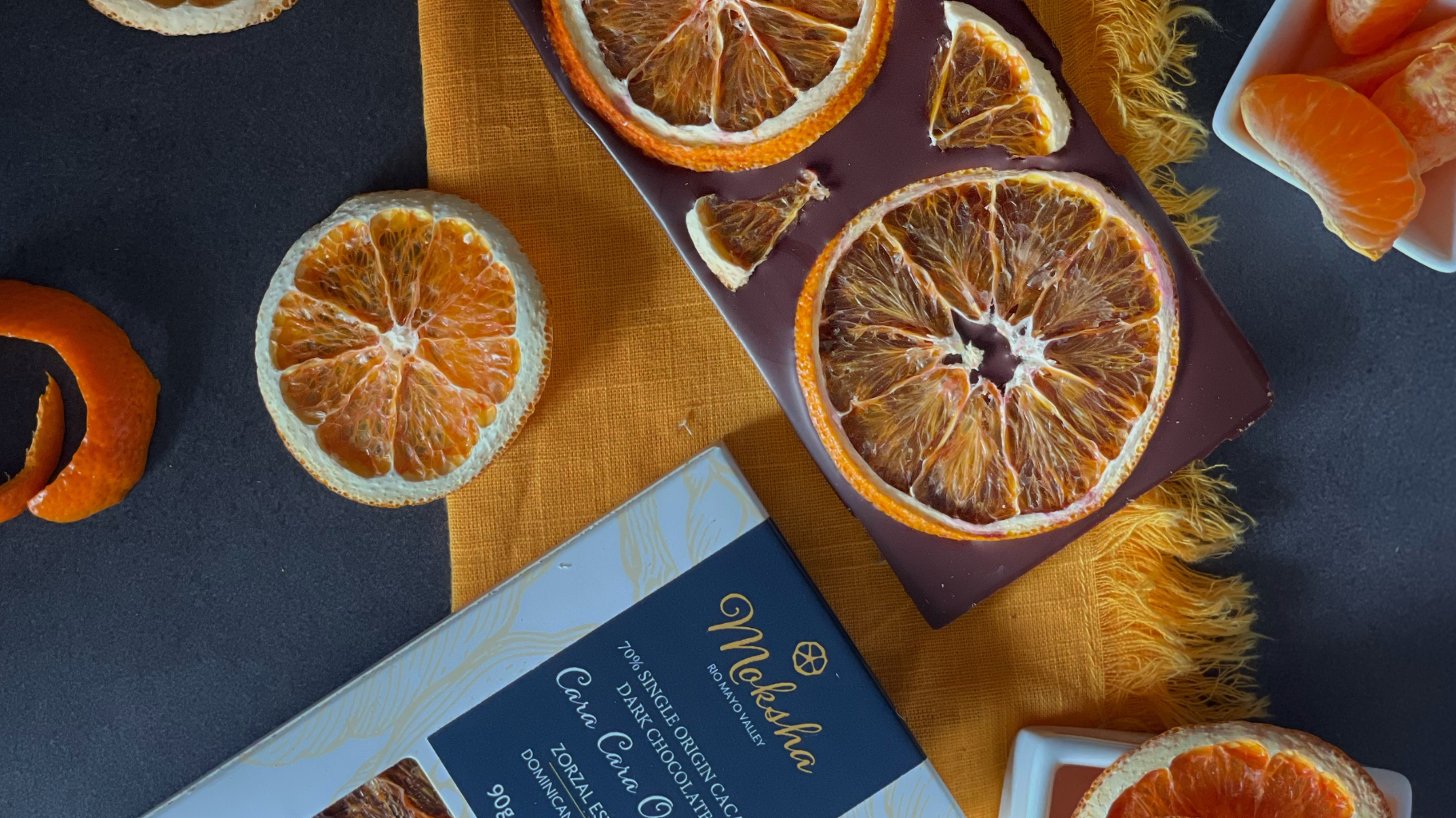 Close-up of Moksha Chocolate’s Cara Cara Orange Dark Chocolate bar topped with dried orange slices, surrounded by orange peels, citrus wedges, and elegant packaging on a dark surface.
