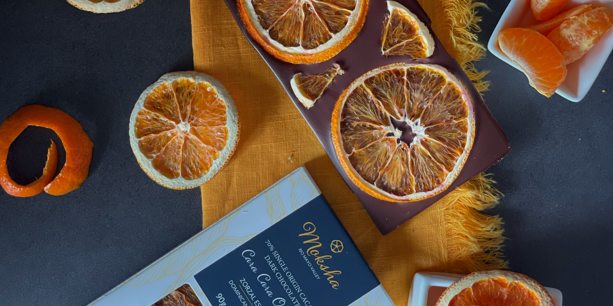 Close-up of Moksha Chocolate’s Cara Cara Orange Dark Chocolate bar topped with dried orange slices, surrounded by orange peels, citrus wedges, and elegant packaging on a dark surface.