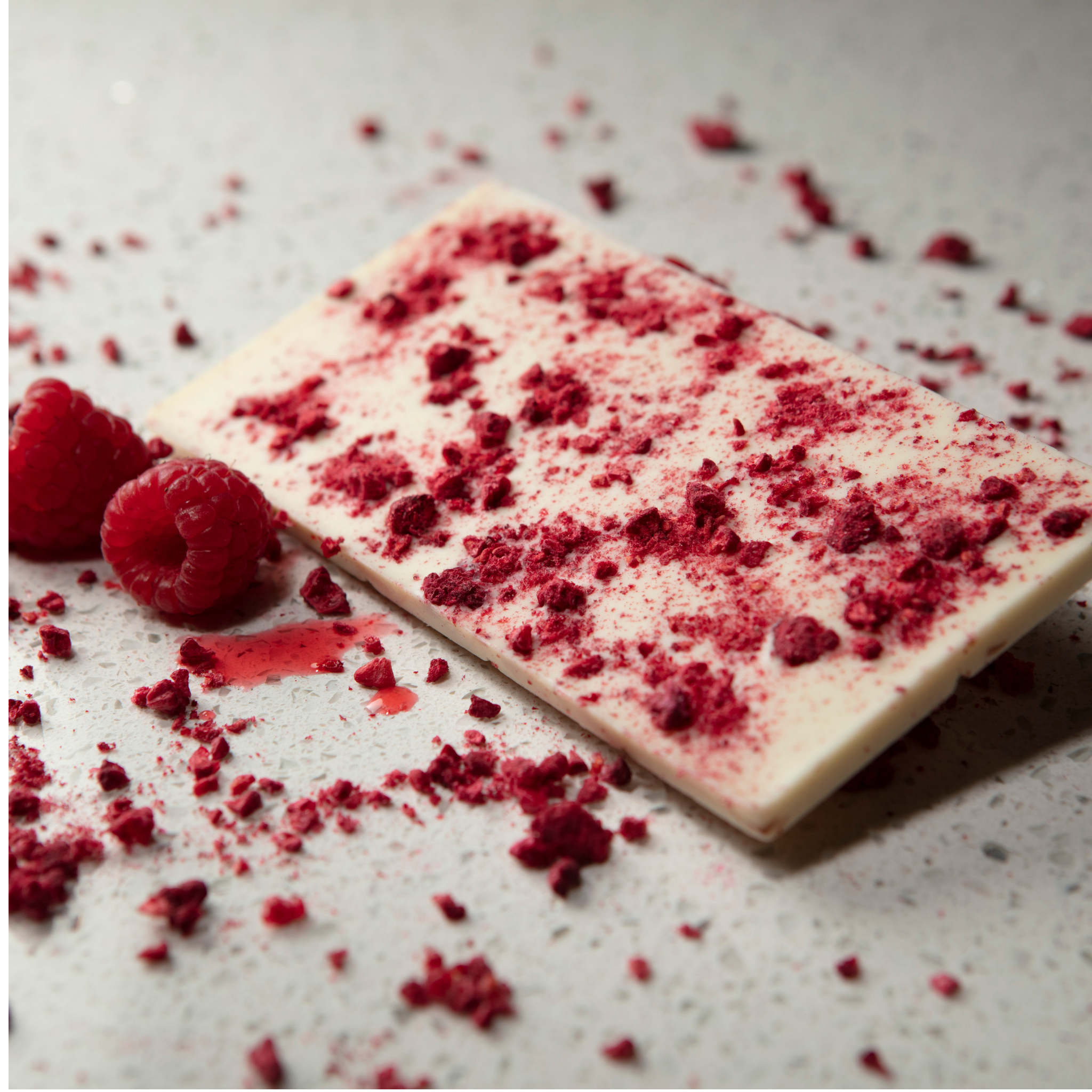 Close-up of Moksha’s vegan white coconut chocolate bar with freeze-dried raspberries, handcrafted with Peruvian cacao, surrounded by fresh raspberries on a light stone surface.