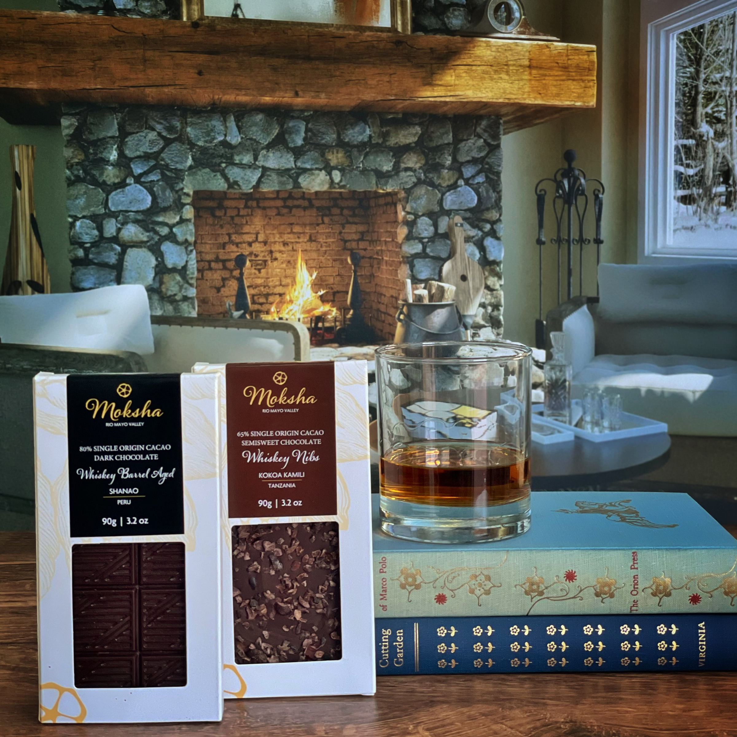 Moksha Chocolate Whiskey Barrel Aged 80% Dark Chocolate and Whiskey Nibs 65% Semisweet Chocolate bars displayed with a glass of whiskey and books in a cozy fireplace winter setting, featuring artisan vegan bean-to-bar chocolate.