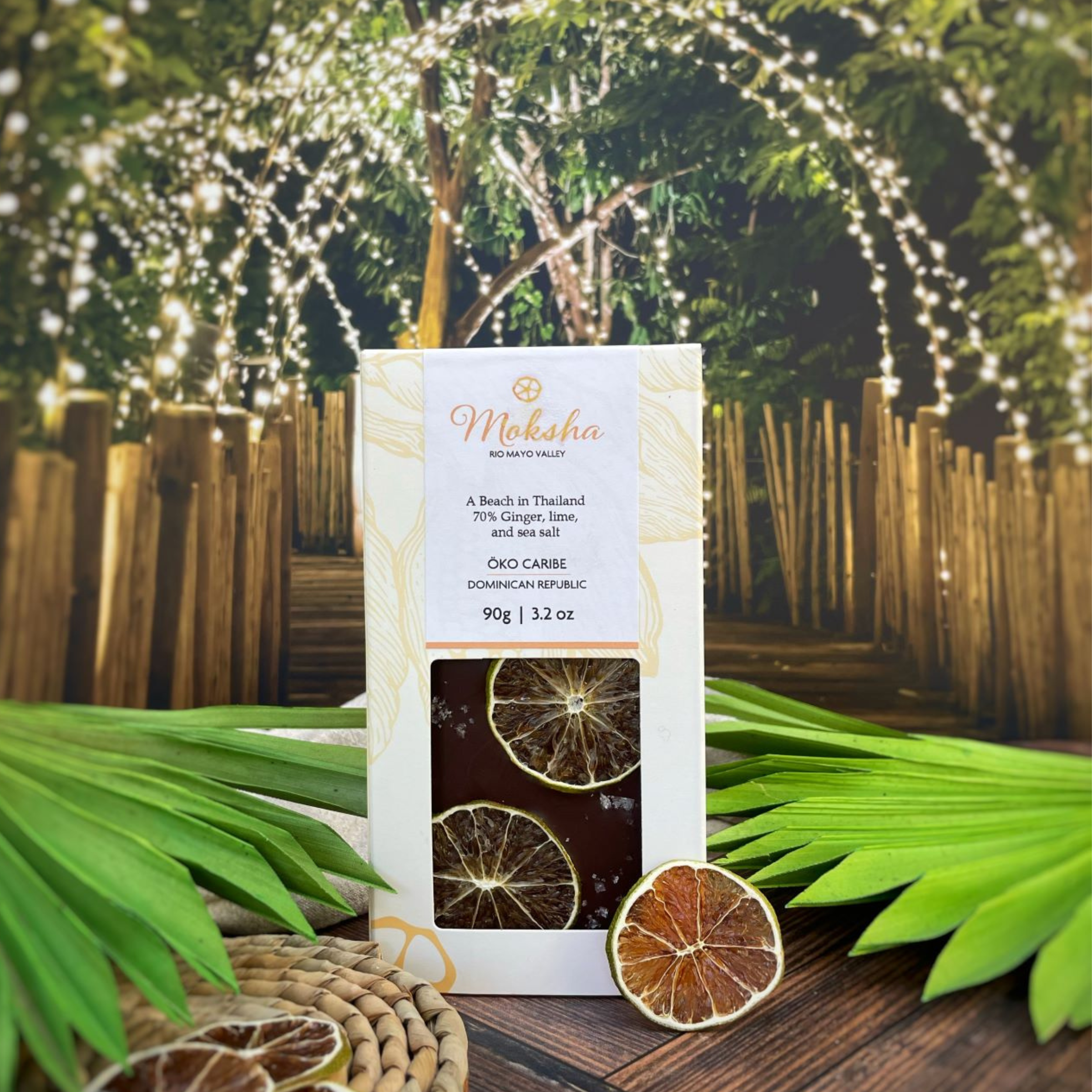 Moksha Chocolate A Beach in Thailand bar featuring 70% dark chocolate with ginger, lime, and sea salt, made with Öko Caribe cacao from the Dominican Republic, displayed with dried lime slices and tropical greenery under twinkling lights.