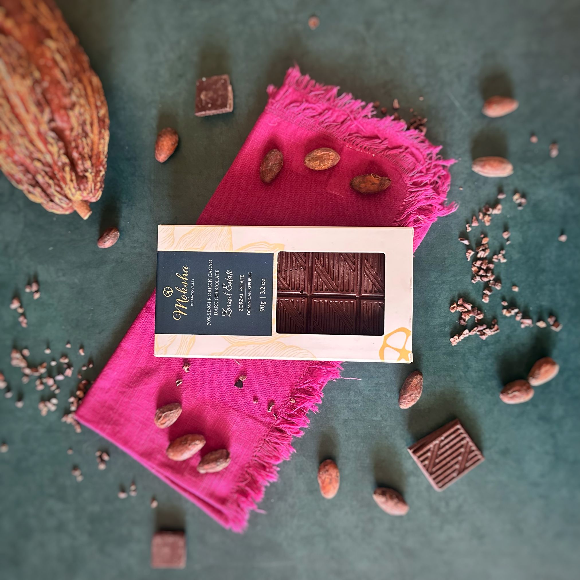 70% dark Zorzal with packaging on a pink cloth surrounded by cocoa beans and a cacao pod.