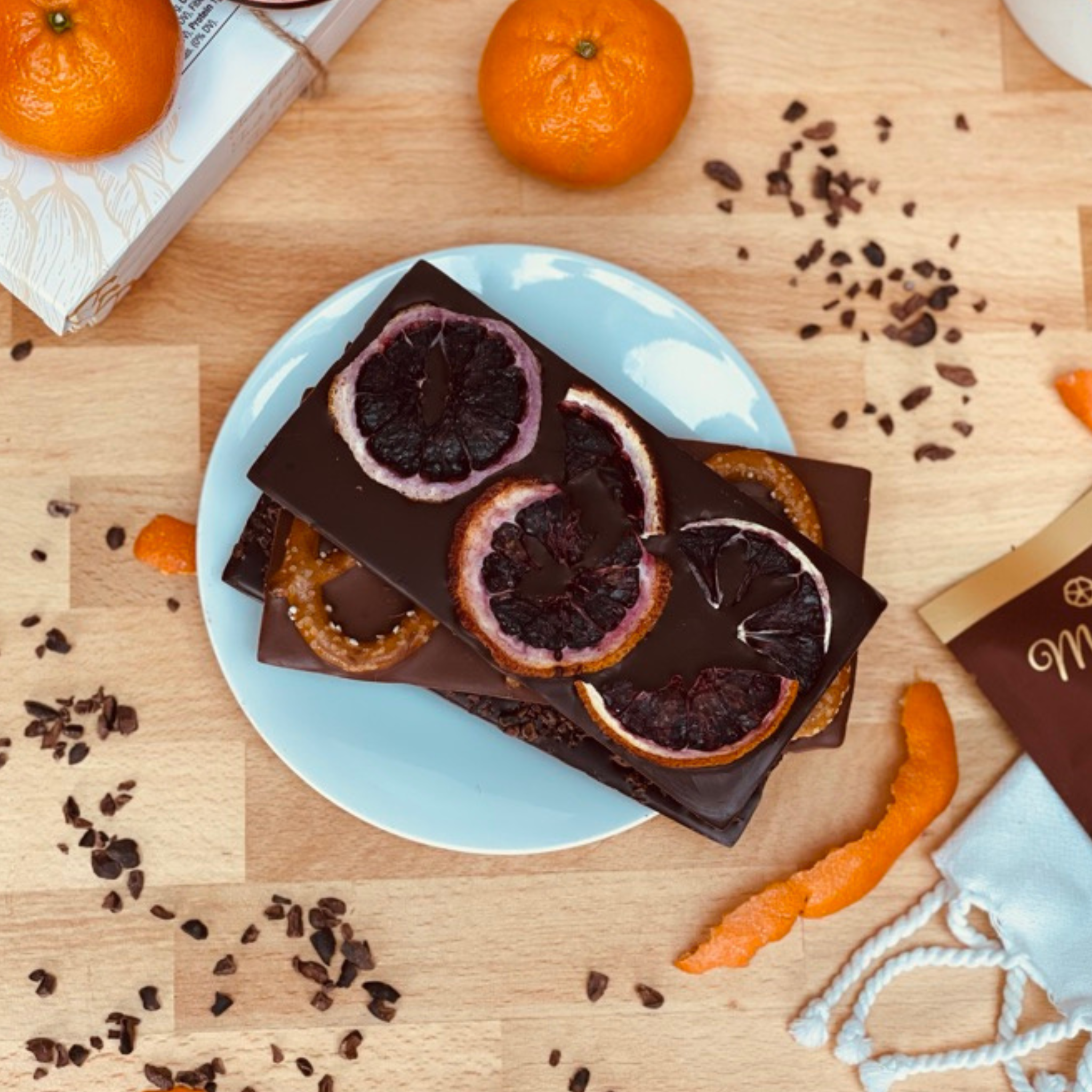 Dark chocolate bars with blood orange slices on a light blue plate, surrounded by oranges and chocolate pieces on a wooden surface.