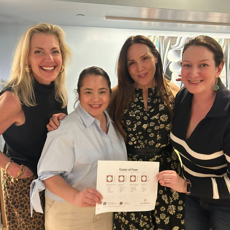 Four women posing together, with one holding a game sheet indoors.
