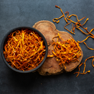 Dried cordyceps militaris mushrooms in a black bowl and scattered on wood, used in natural energy supplements and functional chocolate.