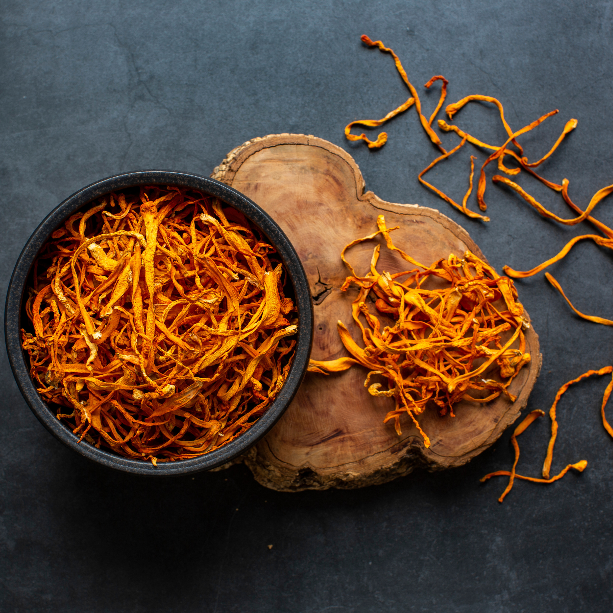 Dried cordyceps militaris mushrooms in a black bowl and scattered on wood, used in natural energy supplements and functional chocolate.