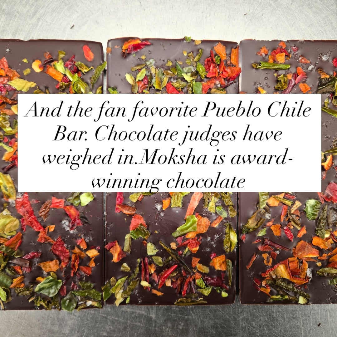 Three rectangular dark chocolate bars topped with colorful dried chili and herb flakes, arranged on a gray surface with a white text overlay reading, 'And the fan favorite Pueblo Chile Bar: Chocolate judges have weighed in. Moksha is award-winning chocolate.