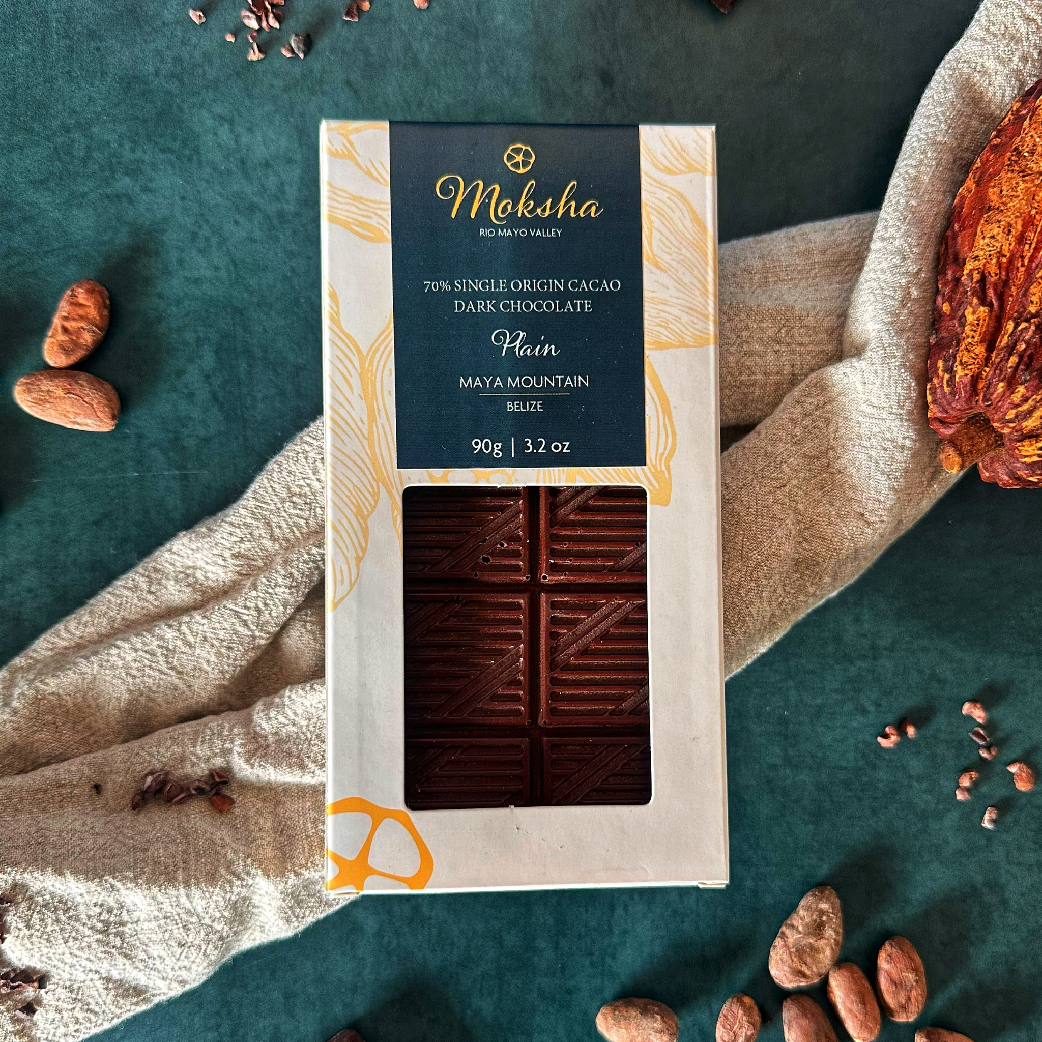 Moksha Chocolate 70% Single Origin Cacao Dark Chocolate bar from Maya Mountain, Belize, in elegant packaging. The bar is partially visible through a window in the box, surrounded by cacao beans and a soft linen cloth on a dark green background.