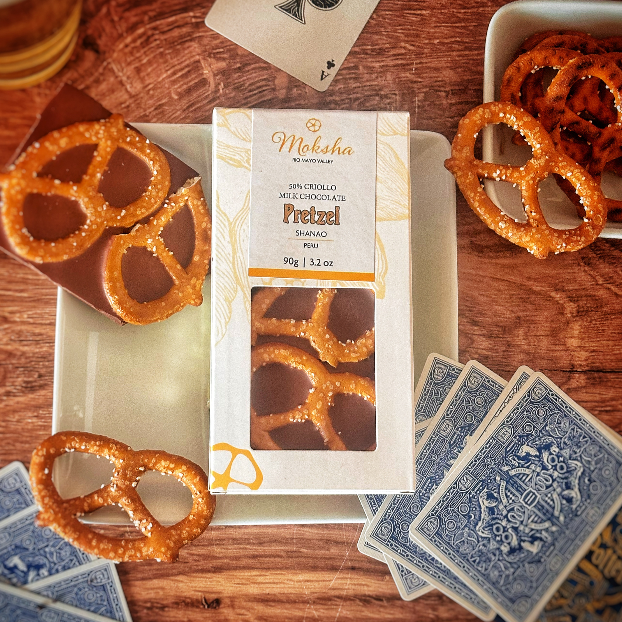 Moksha Chocolate 50% Criollo Milk Chocolate with Pretzel, placed on a plate with a broken piece revealing crunchy pretzel pieces inside. The scene includes a bowl of pretzels, a glass of beer, and playing cards on a rustic wooden table, evoking a cozy and indulgent atmosphere.