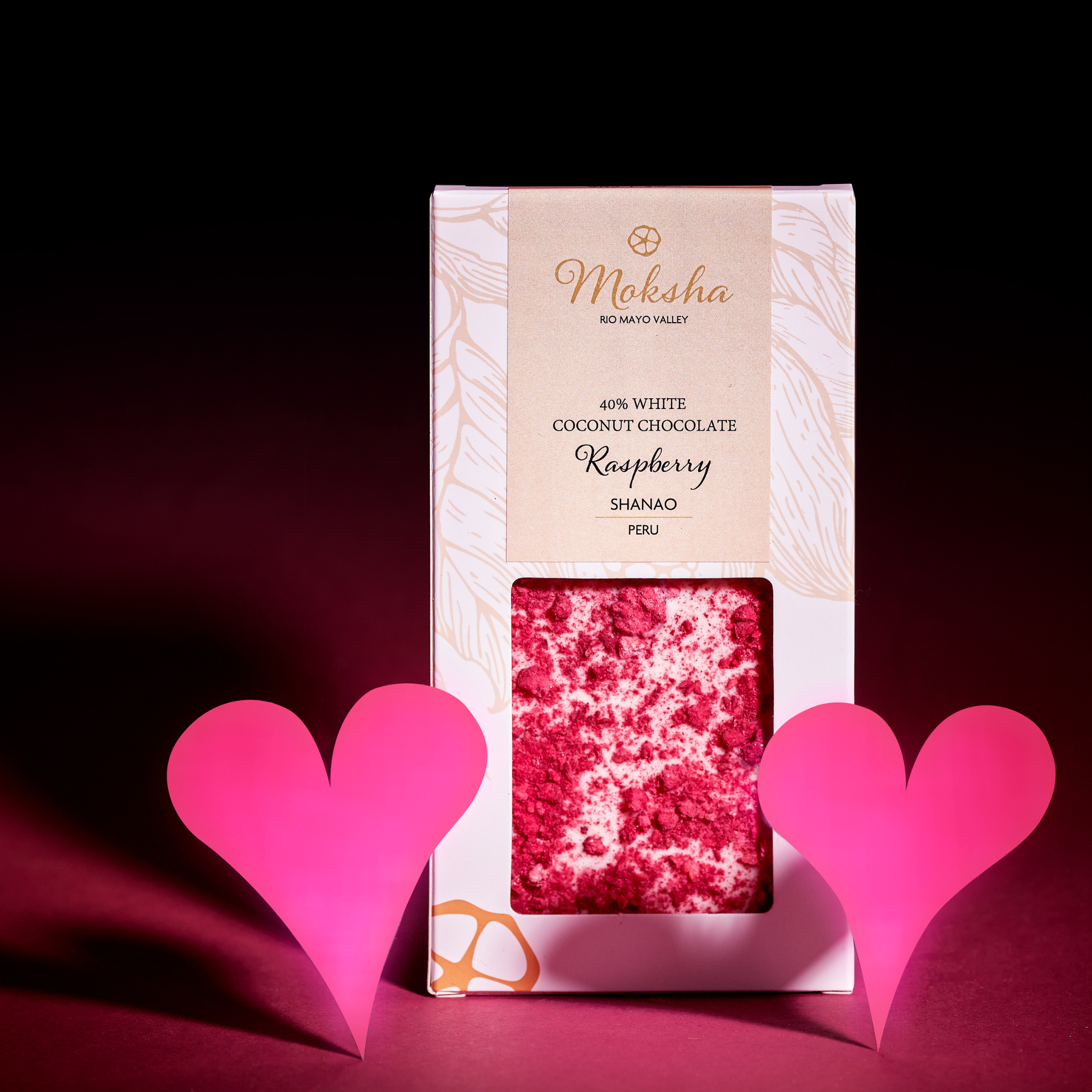 Moksha 40% vegan white coconut chocolate bar with raspberry, made with single-origin cacao from Shanao, Peru, displayed in gift-ready packaging with pink heart decorations on a romantic red background.