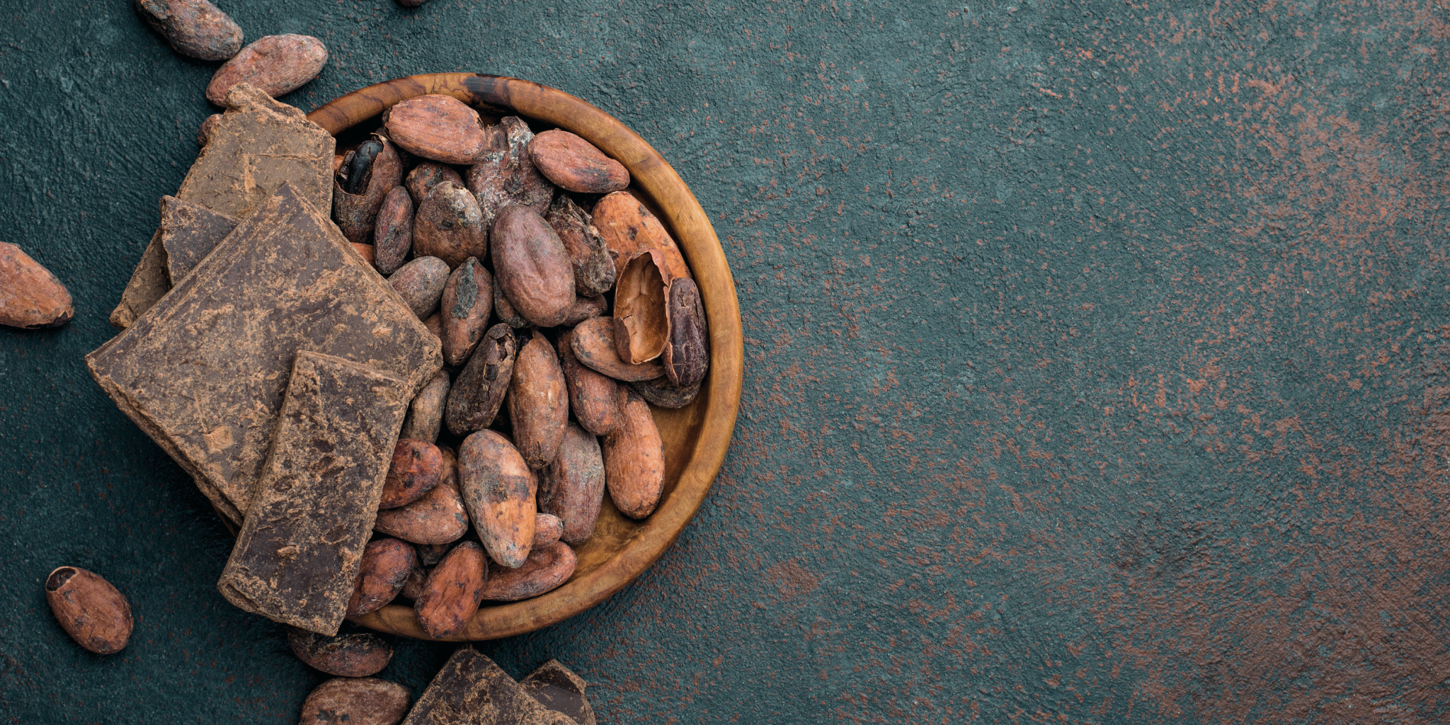 Top-down view of a wooden bowl filled with cacao beans and cacao paste chunks on a textured dark countertop, illustrating raw cacao ingredients.