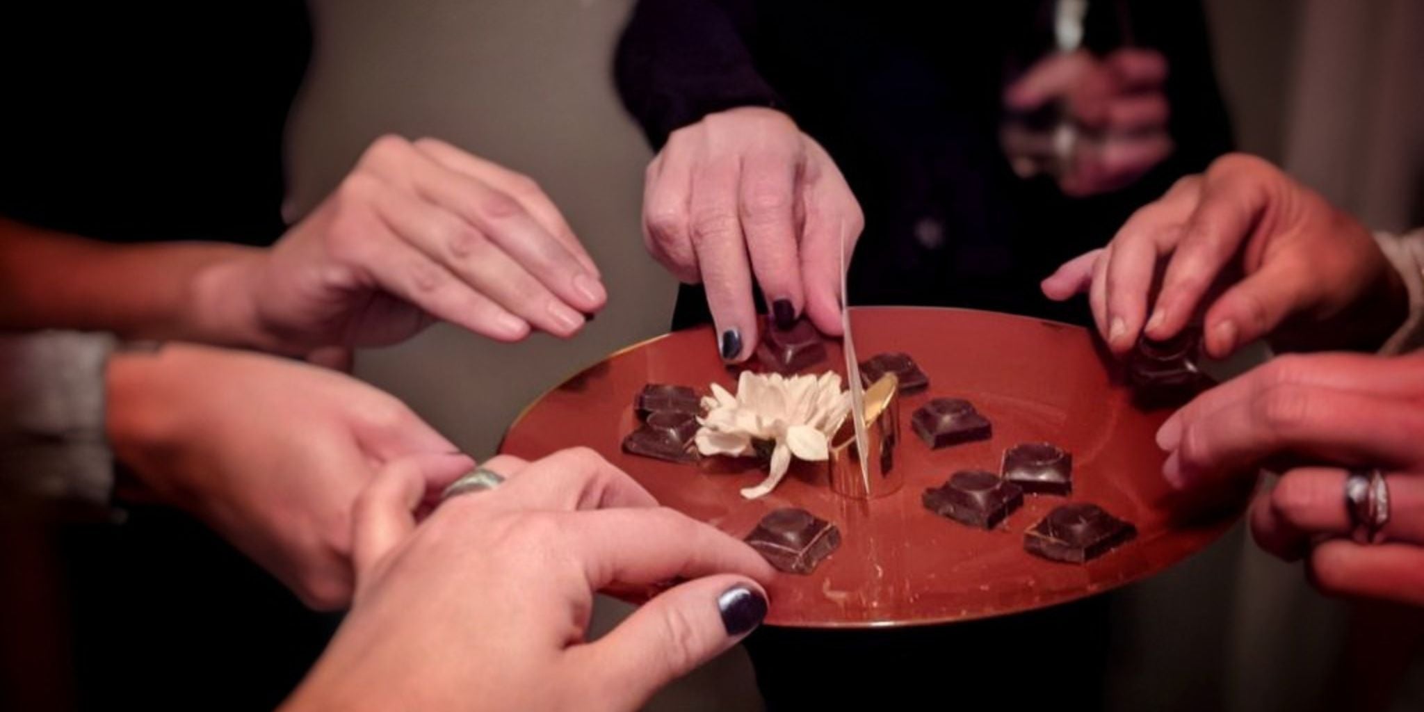 Craft chocolate tasting with hands reaching for dark chocolate squares on a copper platter with a white flower centerpiece.