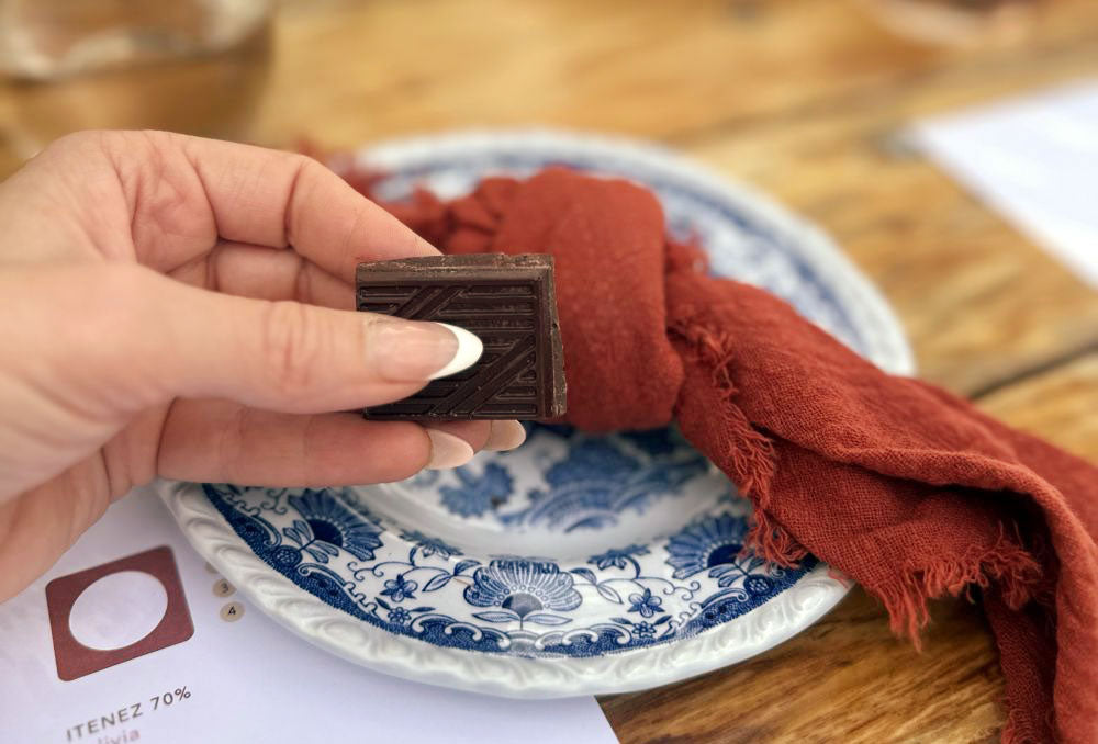 Close-up of a hand holding a square of Itenez 70% dark chocolate over a blue and white patterned plate and rust-colored napkin during a tasting.
