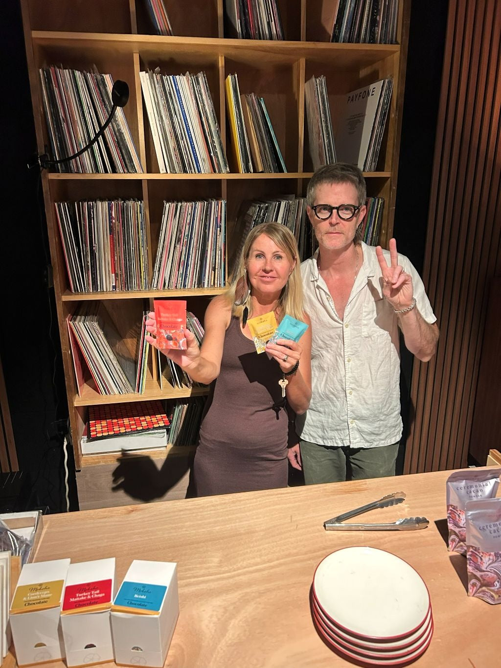 Jen Caines and a man standing behind a wooden counter at a Moksha Chocolate pop-up at FM Denver. Jen is smiling and holding three small Moksha Chocolate packets, with matching chocolate boxes and small plates on the counter in front of a large wooden shelf completely filled with vinyl records.