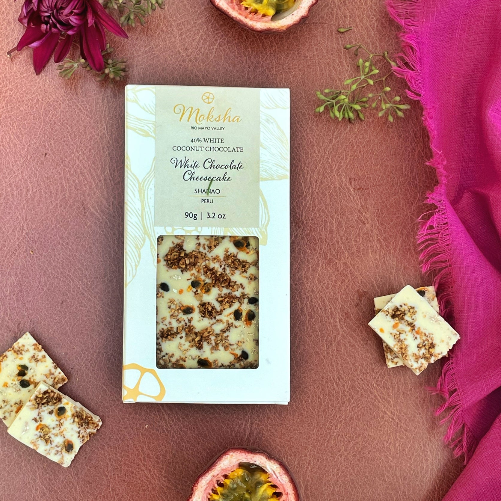 Moksha white chocolate cheesecake bar packaging on a pink surface with decorative elements.