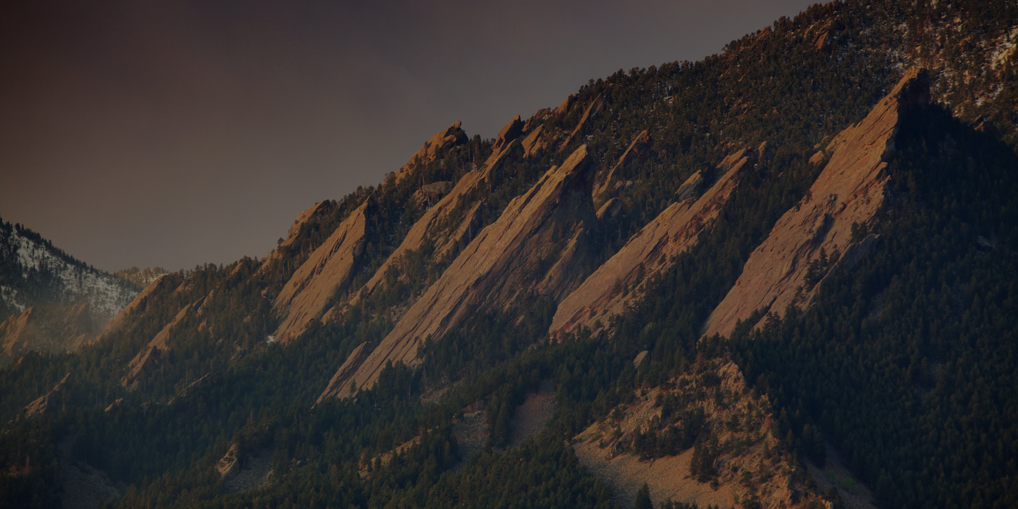 A scenic view of the Flatirons rock formations in Boulder, Colorado, with rugged, slanted sandstone peaks rising against a backdrop of evergreen trees and a dusky sky.
