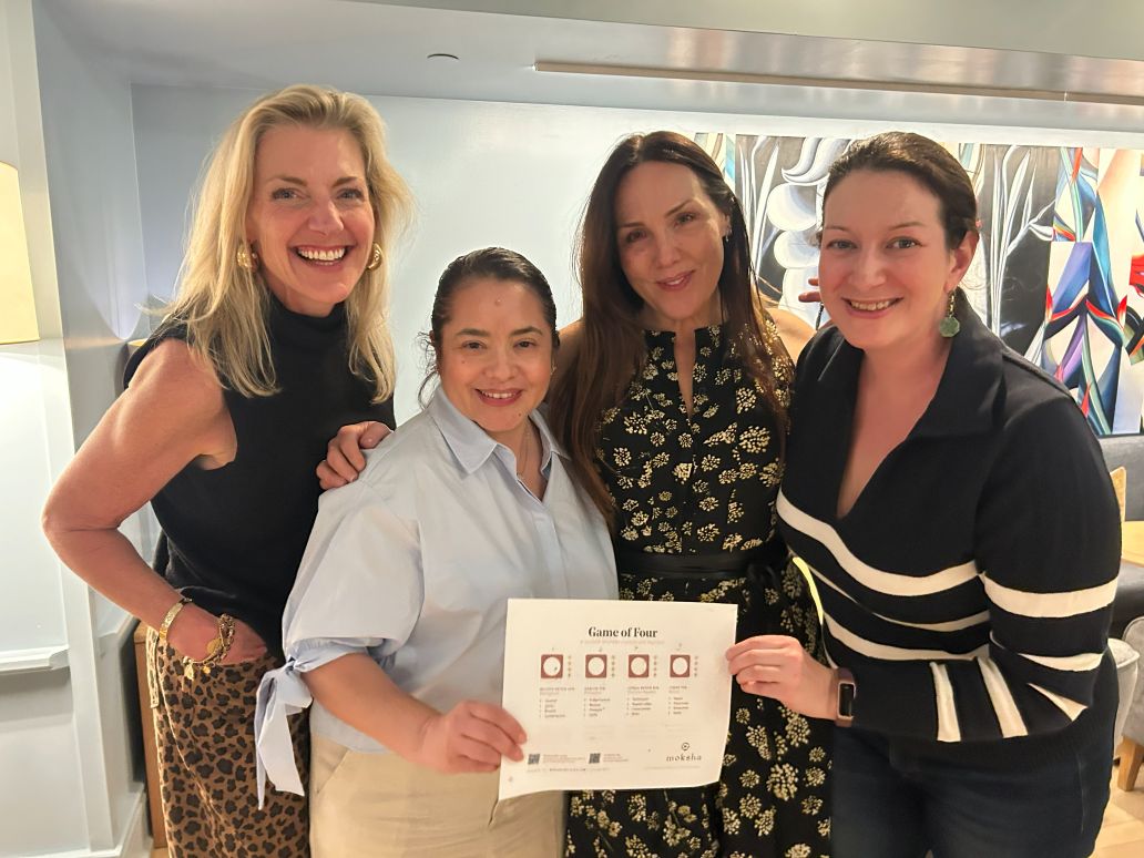 Four women posing together, with one holding a game sheet indoors.