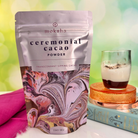 Package of Moksha Ceremonial Cacao Powder (226g/8oz) with vibrant marbled art. A glass of cacao sits beside it on a colorful background—perfect for ceremonial cacao rituals.