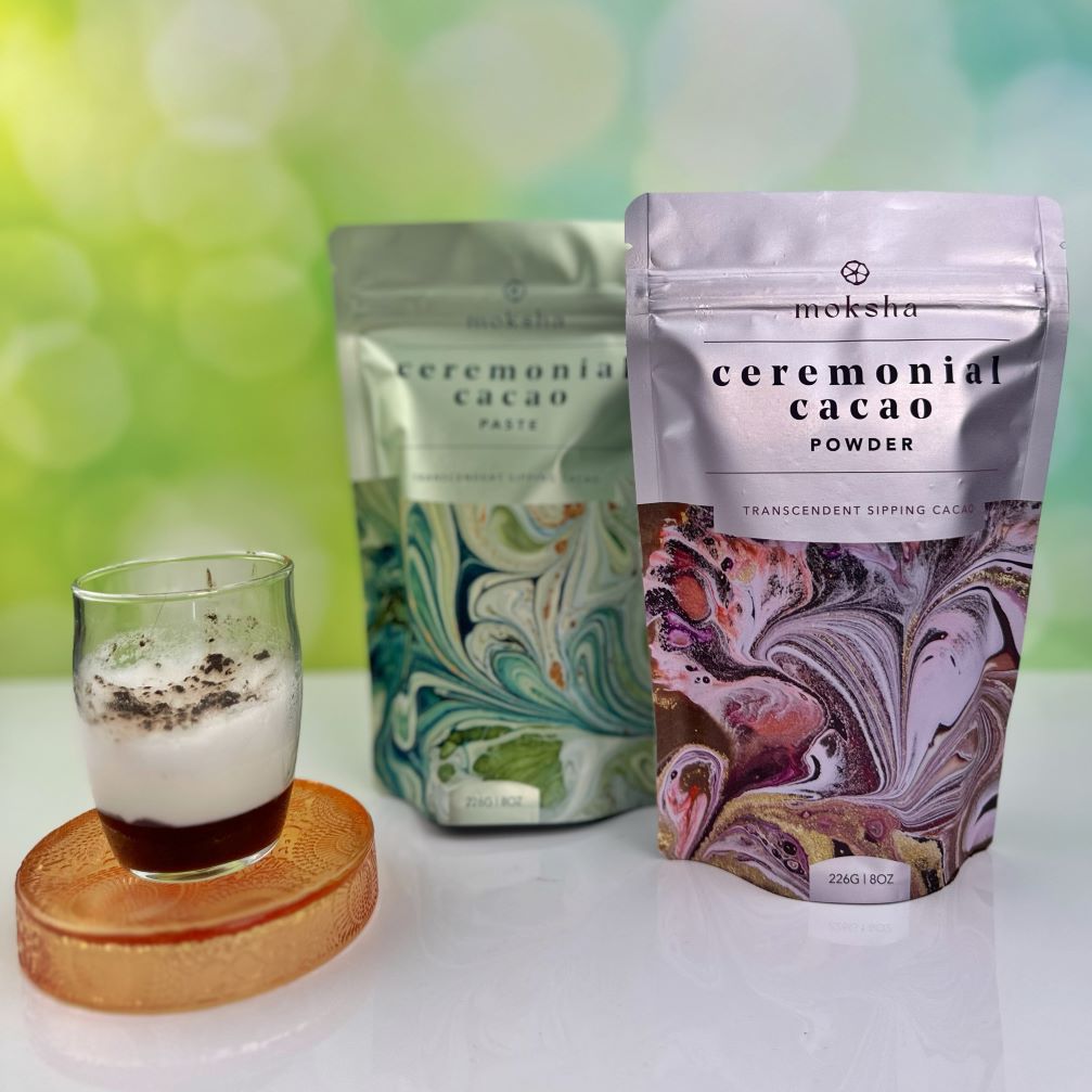 Two packages of ceremonial cacao powder and a glass with a beverage on a white surface with a green blurred background.