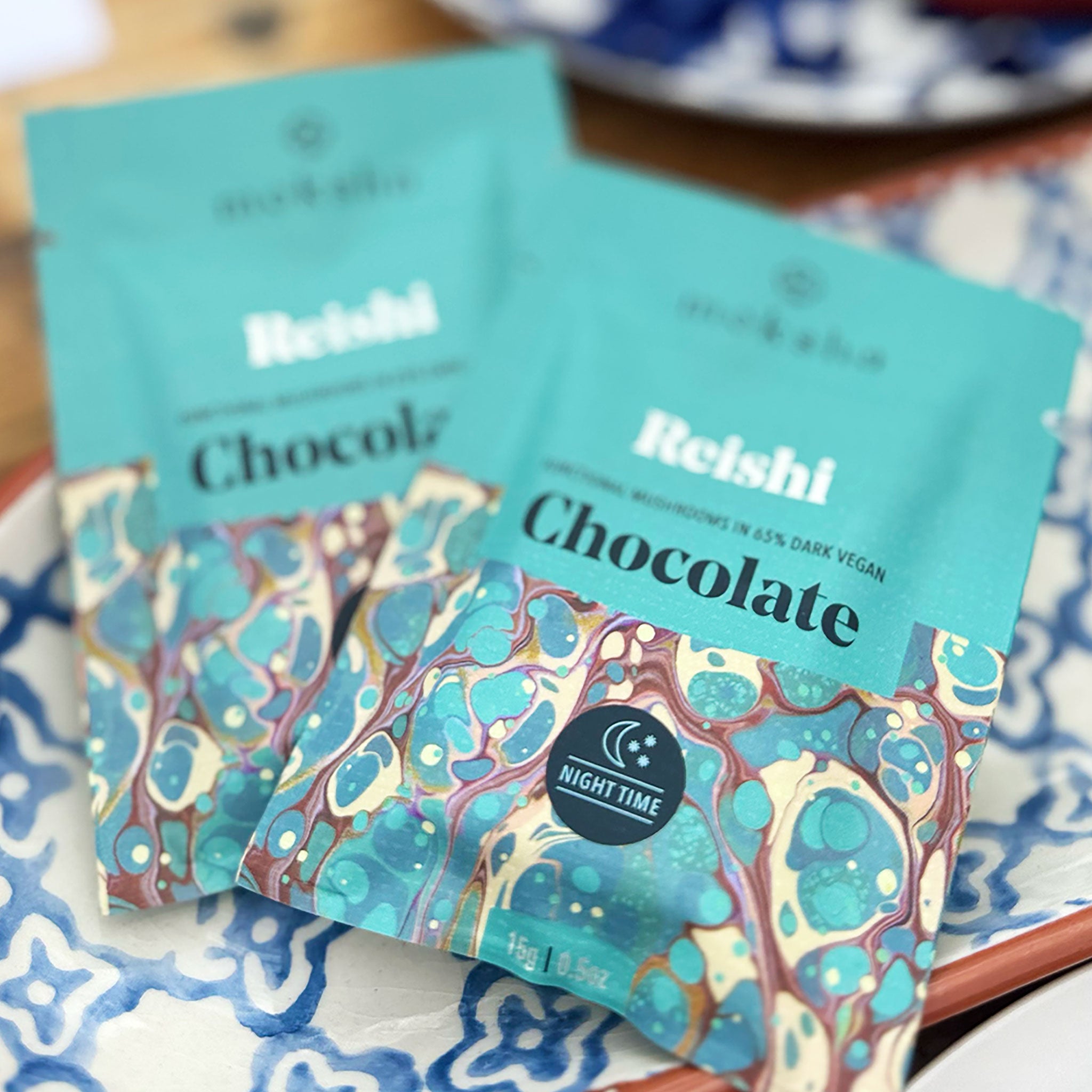 Moksha Reishi mushroom chocolate squares in 65% dark vegan chocolate packaging, shown on a patterned plate with nighttime label and marbled teal design