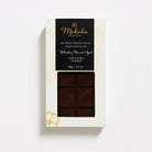 Moksha Whiskey Barrel Aged 80% dark chocolate bar from Kokoa Kamili, Tanzania, in white packaging with a black label and window showing the chocolate pattern.